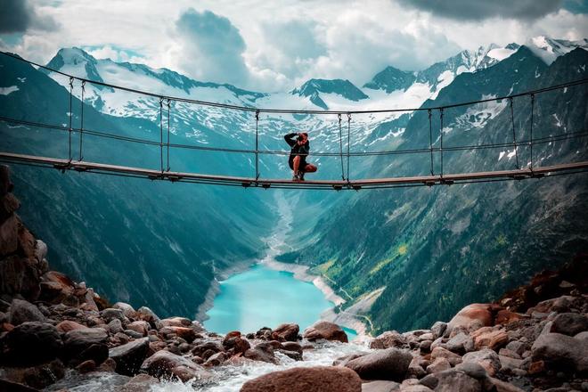 View of mountains and a mountain valey with a lake, a man sitting on a bridge and taking a photo