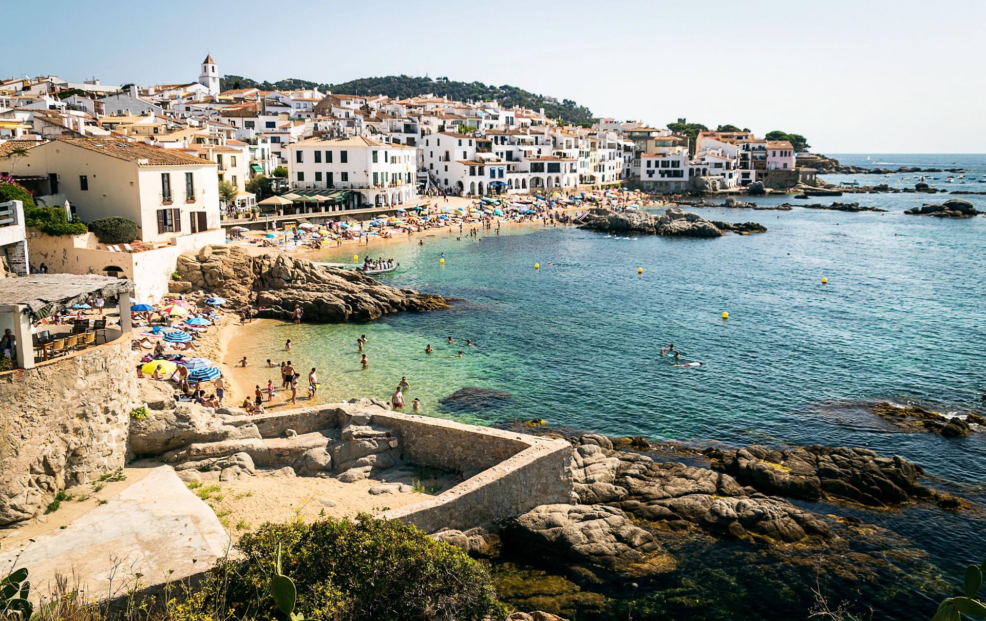 Beach with a lot of people and architecture in Calella de Palafrugell on a day with cloudy weather